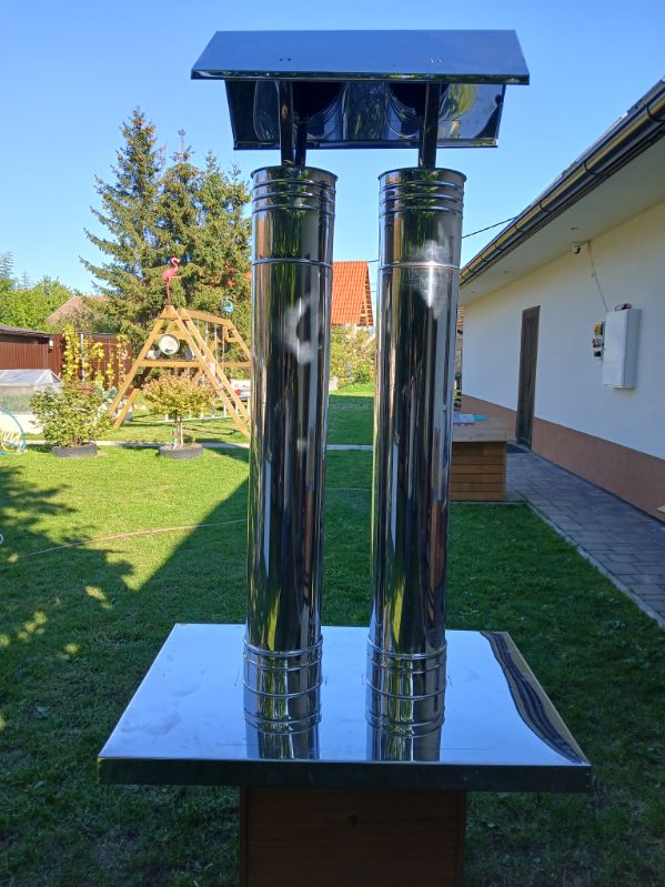 Nerezová strieška na dvojprieduchový komín – dve leštené rúry so spoločnou plochou obdĺžnikovou strieškou na štyroch vzperách, osadené na krycej doske; v pozadí záhrada a rodinný dom